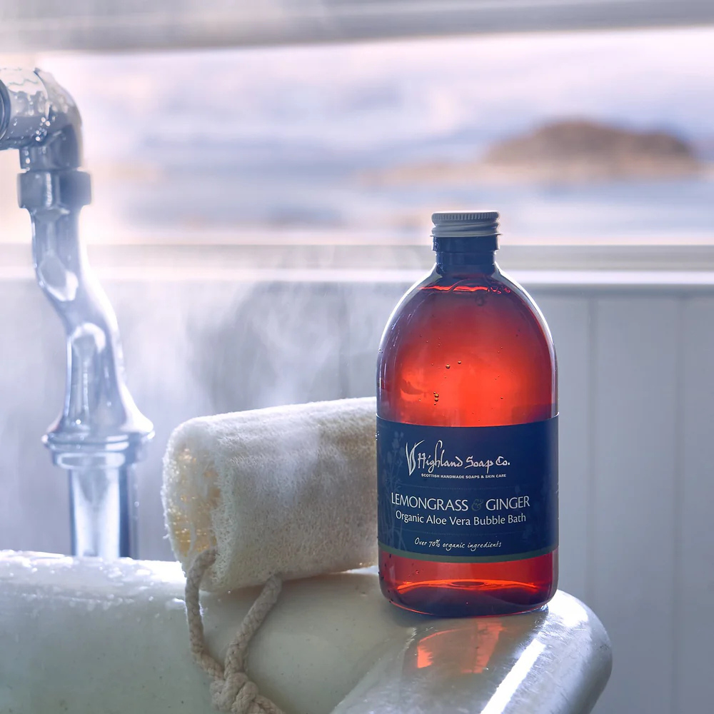 500ml bottle of Lemongrass & Ginger organic bubble bath from The Highland Soap Company, placed beside a candlelit bath with fresh citrus and ginger accents